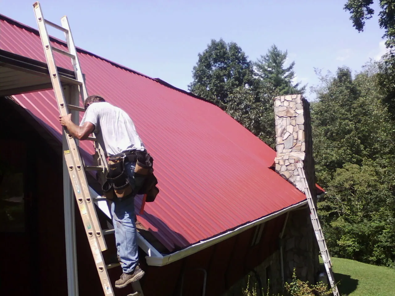 Roofer on a ladder beside a red metal roof and stone chimney — Soffit & Fascia Repair by Roofing Brothers in Elk Creek